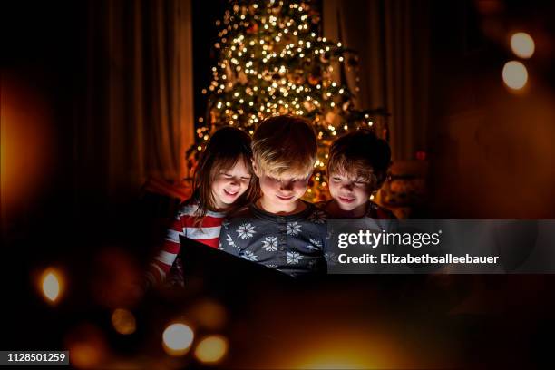 three children sitting in front of a christmas tree reading a book - christmas atmosphere stock pictures, royalty-free photos & images