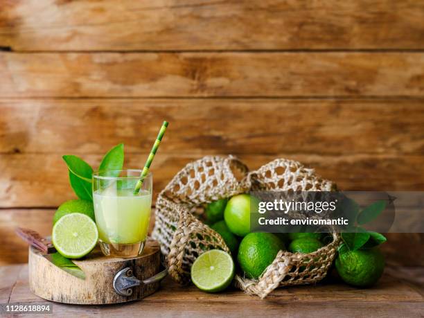 group of limes next to a drinking glass with lime juice and a paper drinking straw on a cutting board with a sharp knife, some limes are cut in cross section and some in a reusable string bag, set against an old weathered wooden panel wall background. - lime juice stock pictures, royalty-free photos & images