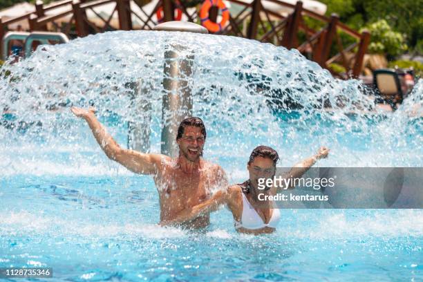 gelukkige jonge paar plezier in zwembad onder waterfontein spatten tijdens vakantie - buitenbad stockfoto's en -beelden