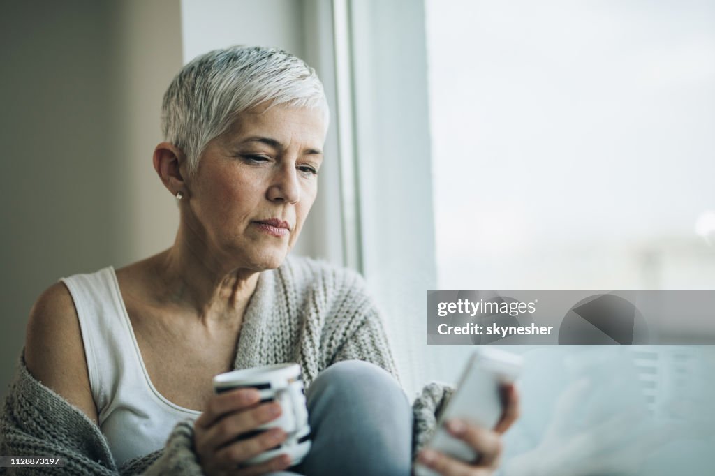 Pensive mature woman reading text message on cell phone by the window.
