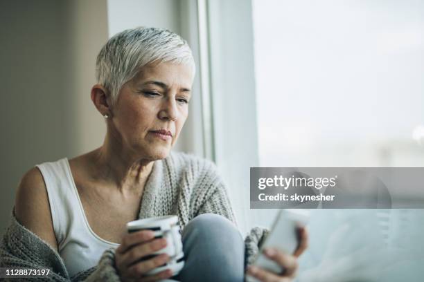 mulher madura pensativa, lendo a mensagem de texto no telefone celular pela janela. - silver surfer - fotografias e filmes do acervo