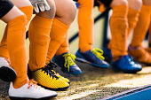 Footballers in soccer cleats. Youth athletes in soccer clothes. Young football players wearing football clothes and soccer shoes sitting on bench in a row. Soccer detail background.