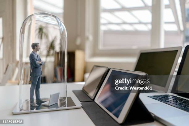 businessman figurine standing under glass bell on desk, facing mobile devices - glass figurine stock pictures, royalty-free photos & images