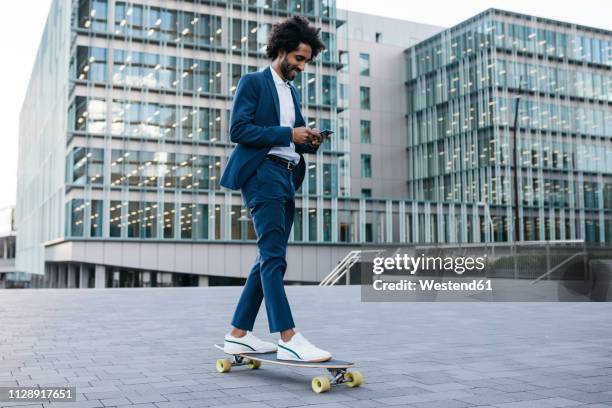 spain, barcelona, young businessman riding skateboard and using cell phone in the city - bien vestido fotografías e imágenes de stock