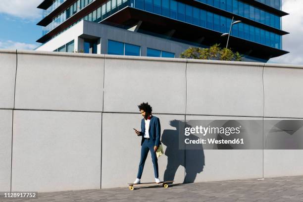 spain, barcelona, young businessman riding skateboard and using cell phone in the city - european outdoor urban walls stockfoto's en -beelden