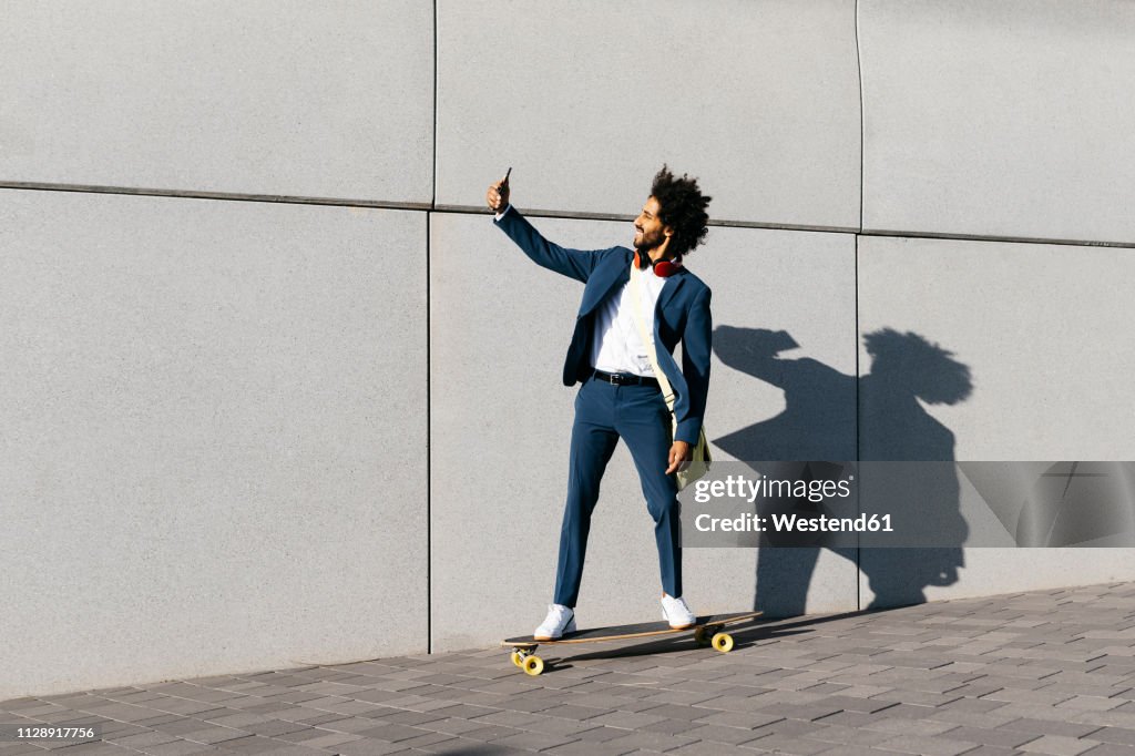 Young businessman riding skateboard along a wall taking a selfie