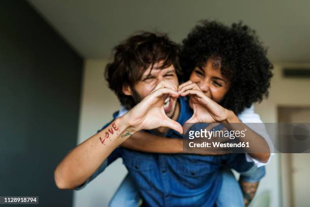 cheerful man carrying girlfriend piggyback at home shaping heart with his hands - caricia fotografías e imágenes de stock