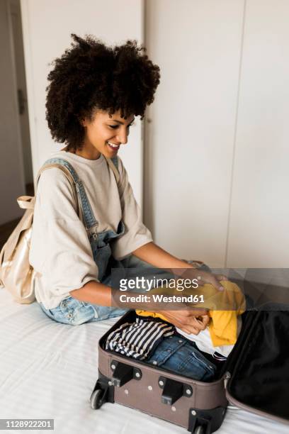 smiling woman sitting on bed with suitcase - packing suitcase stock-fotos und bilder