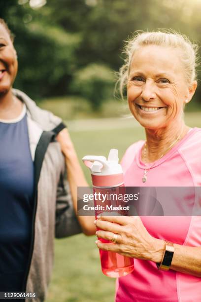 portrait of sporty senior woman holding water bottle while standing with female friend in park - mature woman in water stock pictures, royalty-free photos & images