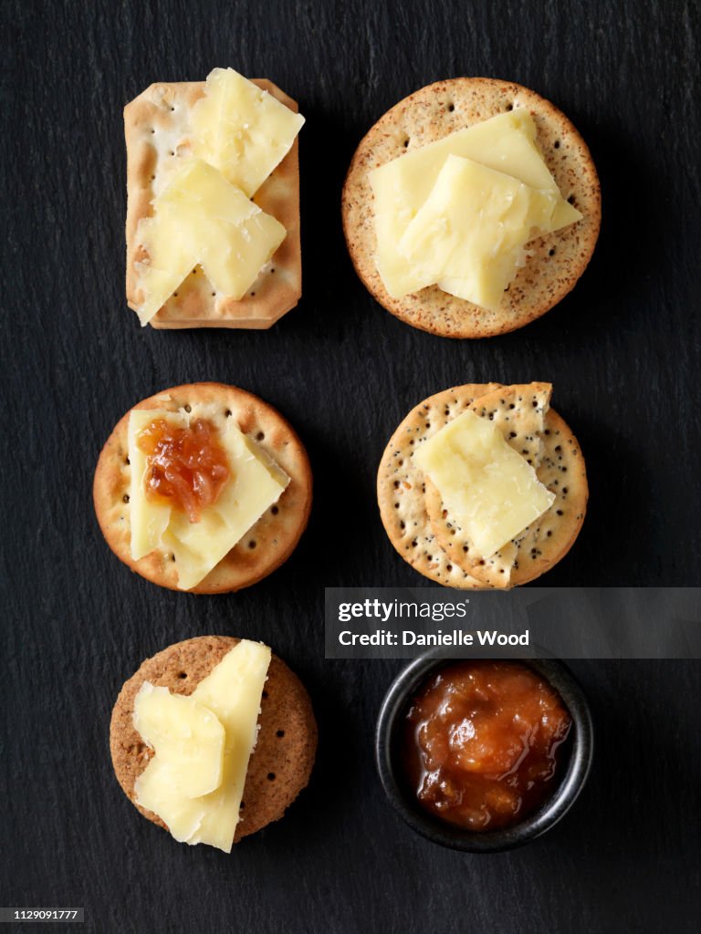 Still life of variety of cheese crackers with cheddar cheese and chutney on black slate, overhead view