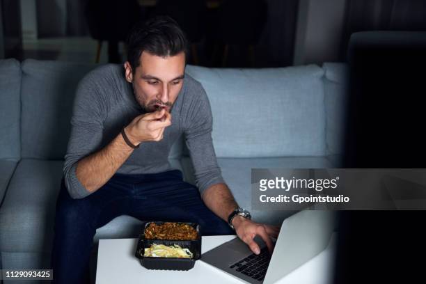 young man sitting on sofa in evening eating takeaway and using laptop - telecommuting eating stock pictures, royalty-free photos & images