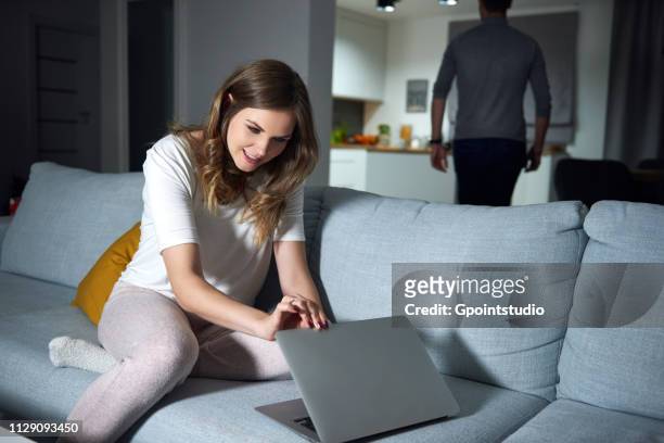 young woman sitting on sofa in evening, peeking at boyfriend's laptop - entrometido fotografías e imágenes de stock
