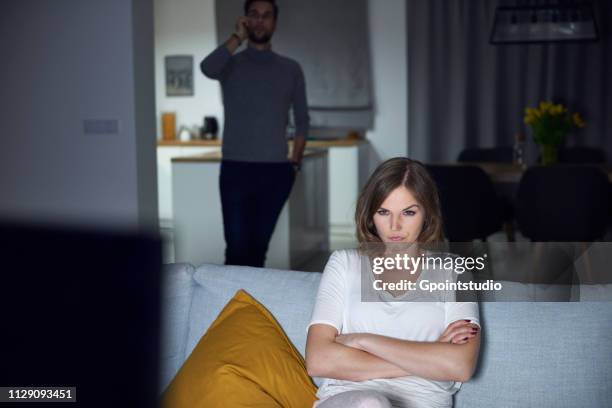 young woman sulking on sofa in evening, while boyfriend makes smartphone call - neid stock-fotos und bilder