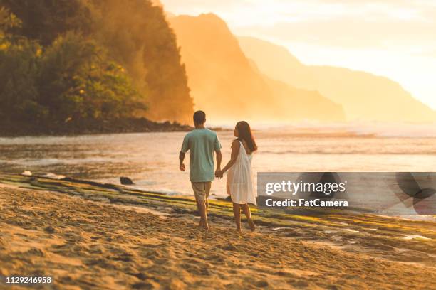 a striking eurasian couple holds hands as they walk along the beach at sunset - kauai stock pictures, royalty-free photos & images