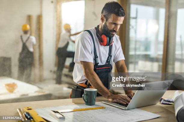 sonriente trabajador manual usando una computadora en sitio de construcción. - building contractor fotografías e imágenes de stock