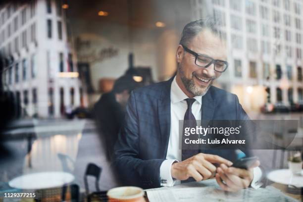 mature businessman sitting inside at cafe and using phone - homme d'affaires photos et images de collection