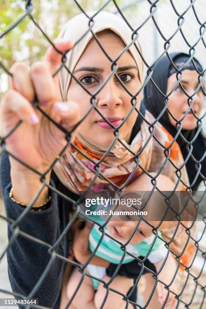 vrouwelijke moslim vluchtelingen holding haar baby kijken door een hek - syrian refugees stockfoto's en -beelden