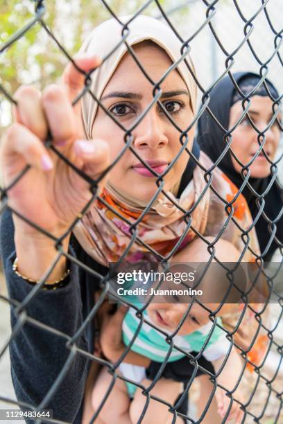 female muslim refugee holding her baby watching through a fence - syrian war stock pictures, royalty-free photos & images