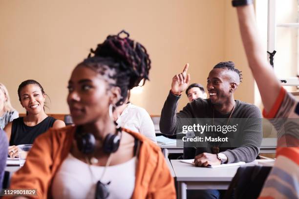 happy young male student raising hand while sitting with classmates in school building - student visa stockfoto's en -beelden