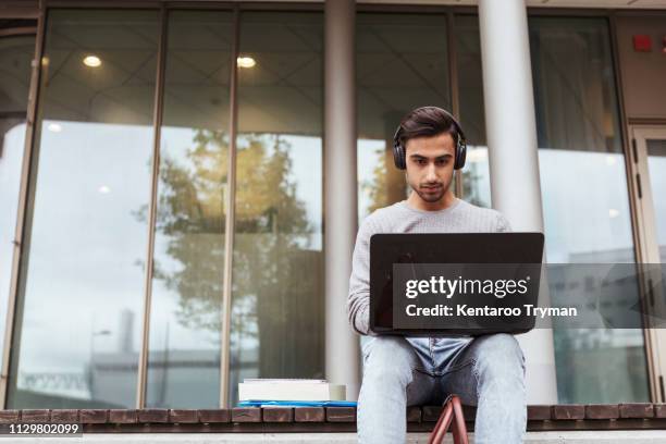 low angle view of university student wearing headphones while using laptop against glass window at campus - bank student stock-fotos und bilder