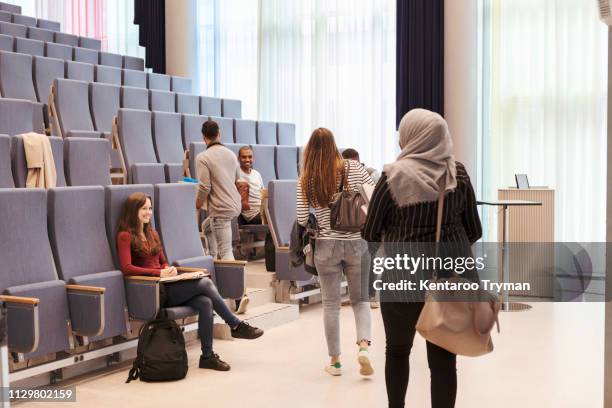 smiling students looking at friends walking towards chairs in lecture hall at high school - religious veil stock pictures, royalty-free photos & images