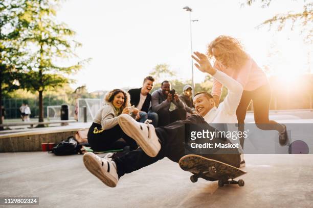 laughing friends photographing man falling from skateboard while woman pushing him at park - active lifestyle stock-fotos und bilder