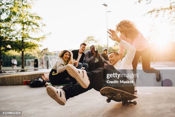 laughing friends photographing man falling from skateboard while woman pushing him at park - distante fotografías e imágenes de stock