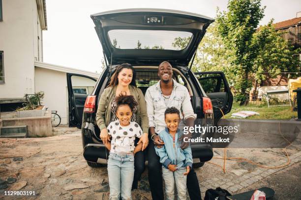 portrait of smiling multi-ethnic family leaning on car trunk in front yard - car in driveway foto e immagini stock