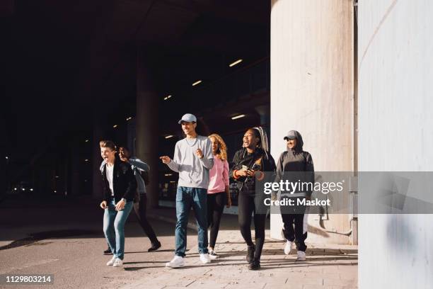 happy friends looking away while walking on street under overpass in city during sunny day - adolescence photos et images de collection