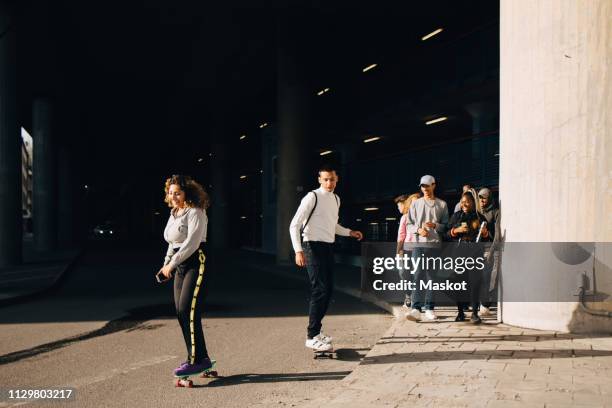 friends looking at man and woman skateboarding on road during sunny day - arab group stockfoto's en -beelden