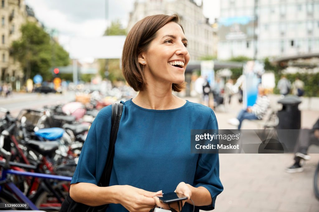 Smiling mid adult businesswoman looking away while holding smart phone in city