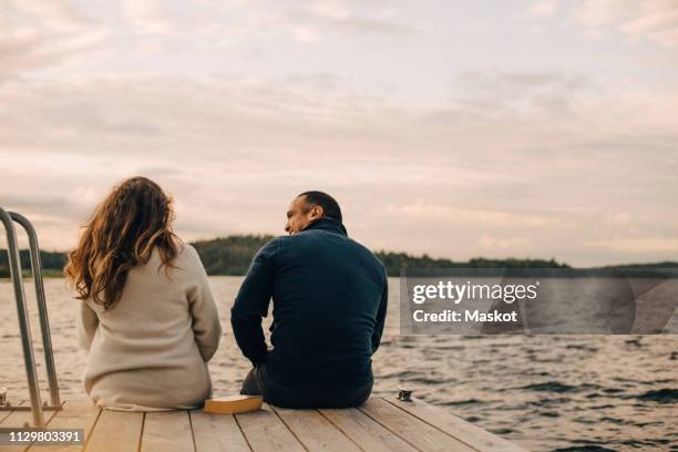 rear view of couple talking while sitting on jetty by lake against sky - cottage exterior stockfoto's en -beelden