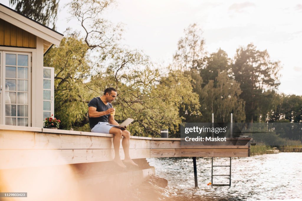Mature man having juice while using laptop on patio by lake