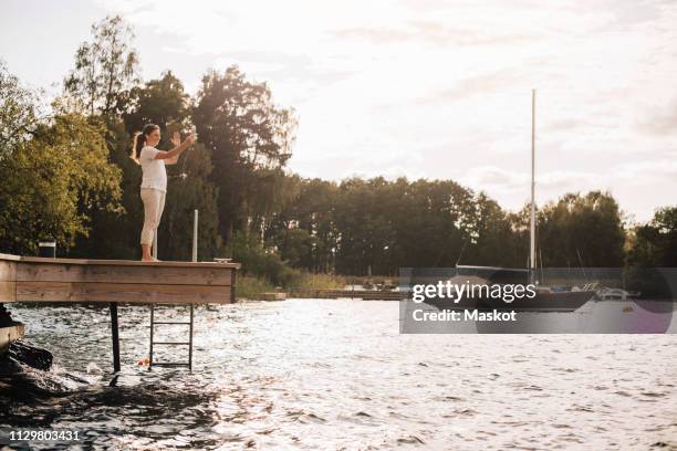 full length of woman talking through video call on mobile phone while standing at patio by lake against sky - voip stock-fotos und bilder