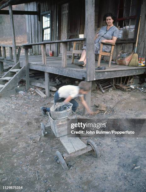 Woman sits on the porch of a log house as a child is playing in front of it with a bucket of coal, Pike County, Kentucky, US, 1967.