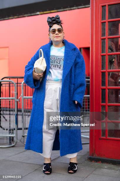 Guest is seen on the street wearing blue coat, white satin pants, tie dye shirt and black hair bow with white bag and black shoes during London...