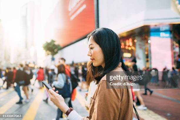 young woman checking on mobile phone while crossing street and commuting in busy downtown city street - downtown city stock pictures, royalty-free photos & images