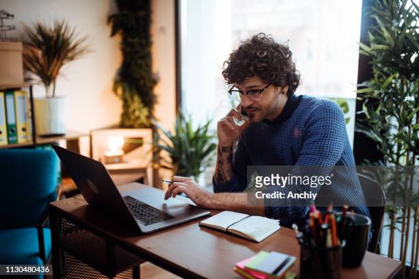hombre joven hablando por teléfono en su oficina en casa - hipster professional fotografías e imágenes de stock