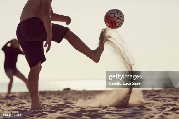 young boy in shorts kicks a football on the beach with sand flying in its wake - kicking sand stock pictures, royalty-free photos & images