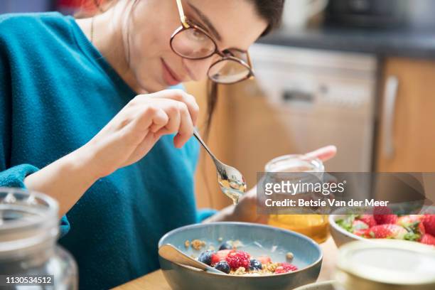 woman pours honey over her breakfast bowl, sitting at kitchen table. - miel photos et images de collection