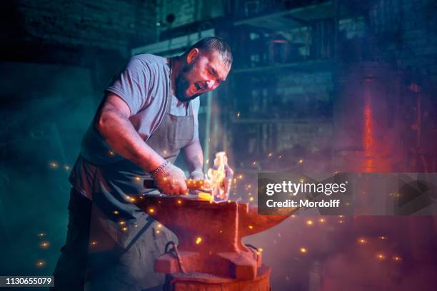 big bearded blacksmith is forging metal rod on the anvil - schmied stock-fotos und bilder