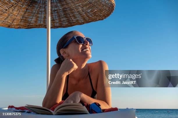 schöne frau liest buch auf dem liegen am strand - sonnenbaden stock-fotos und bilder