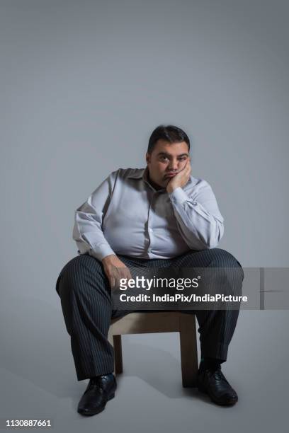overweight man sitting on chair in sad mood with chin resting on hand - man plain background stock pictures, royalty-free photos & images