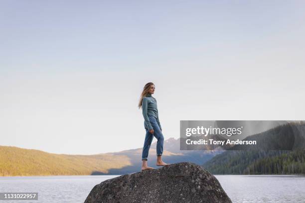 young woman standing on boulder looking at mountain lake - jeans barefoot fotografías e imágenes de stock