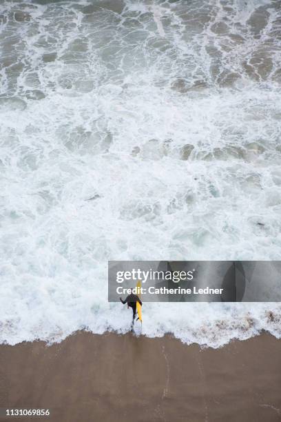 man in black wetsuit running into ocean from above holding yellow surfboard - beach la stock pictures, royalty-free photos & images