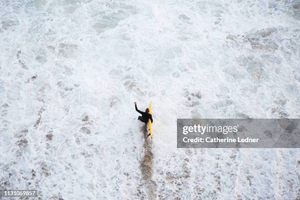 surfer in black wet suit walking into turbulent waters carrying yellow surfboard - californie surf stock pictures, royalty-free photos & images