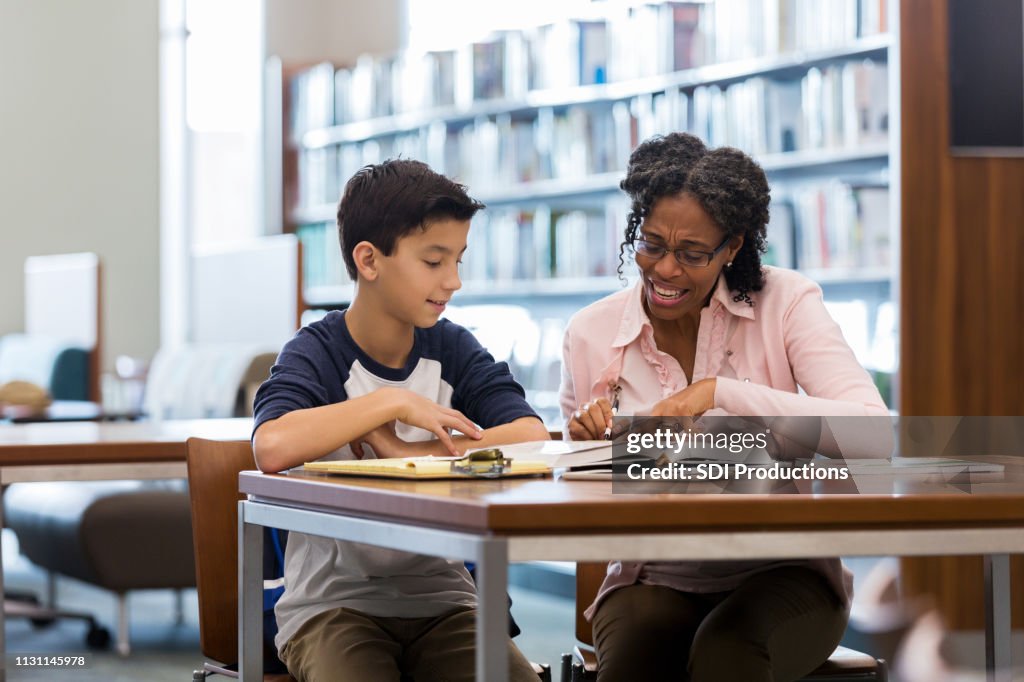 O menino da idade da escola média lê com mentor na biblioteca