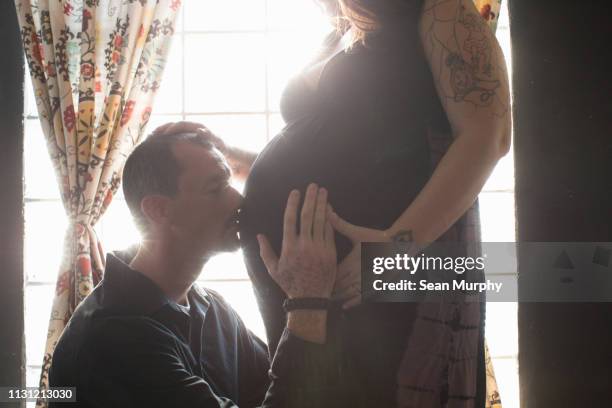 cropped view of tattooed pregnant mid adult woman, partner kneeling kissing stomach - belly kissing stock-fotos und bilder