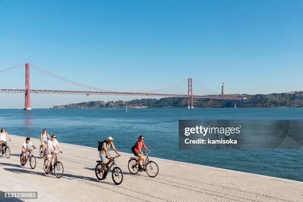 people ride their bicycles along the maat museum. behind it is the bridge of april 25 and christ the king. - tagus river stock pictures, royalty-free photos & images