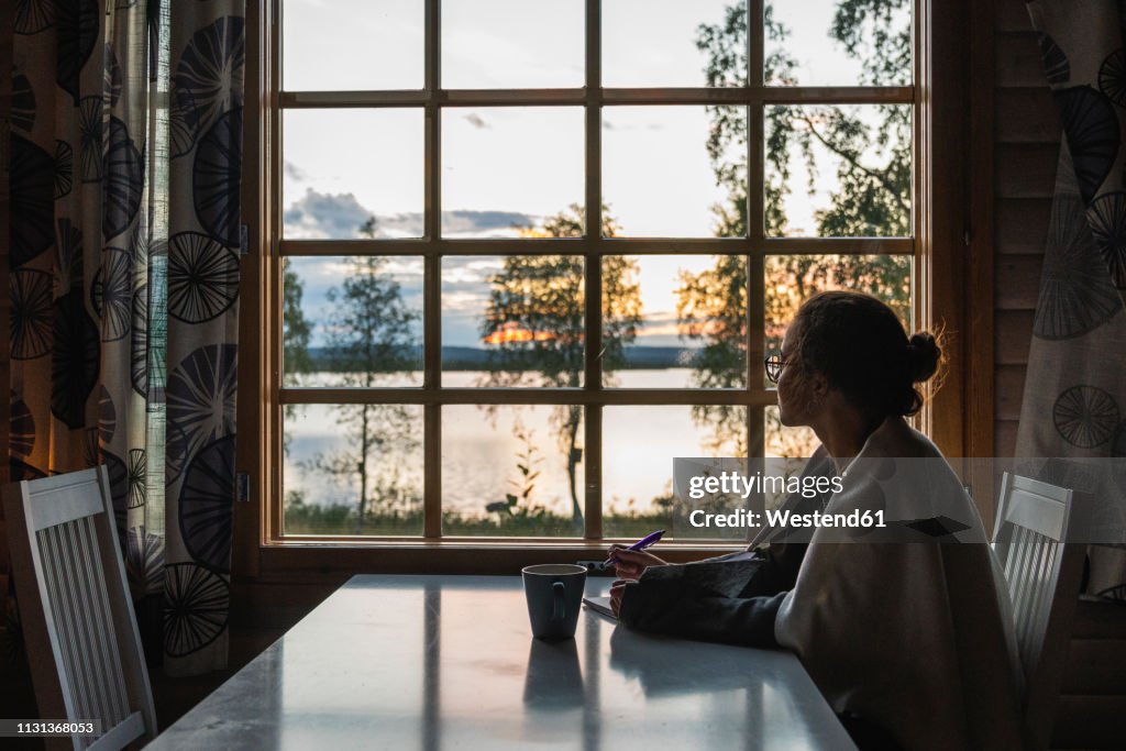 Finland, Lapland, young woman sitting at the window looking at a lake